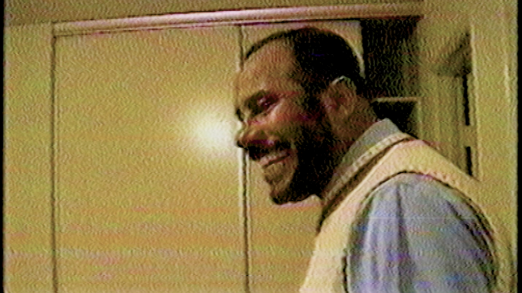 A grainy photo shows a man smiling in profile. There is a beige wall behind him with a storage space alcove. He is wearing a cream colored vest sweater and a light blue longsleeved shirt.
