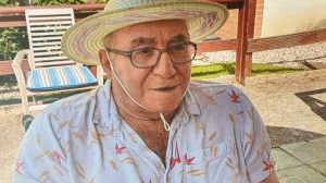 An old man sits in a chair and smiles. He is wearing glasses and has a straw hat on which is tied under his chin. He is wearing an open-necked shirt with a red and yellow pattern on it. He is outside and the sun is shining behind him.
