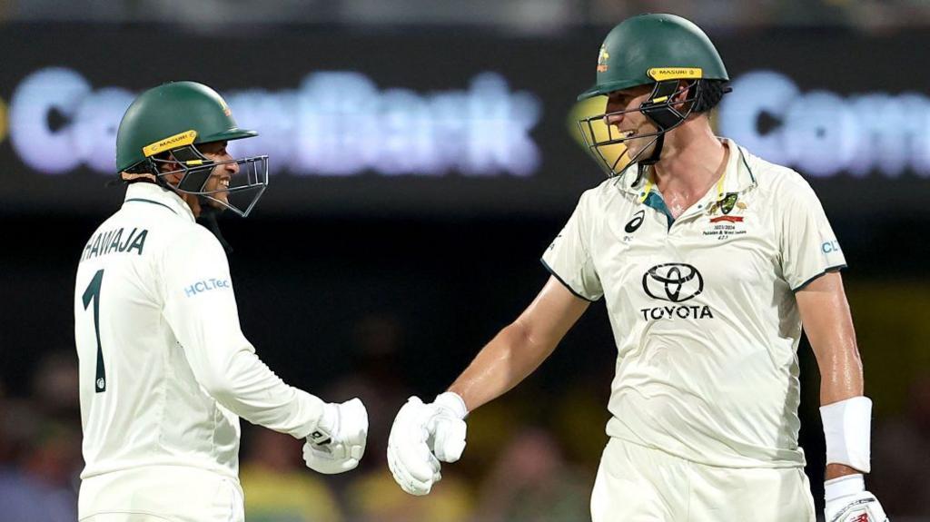 Usman Khawaja and Pat Cummins smile at each other while wearing batting helmets, with their right batting gloves almost touching