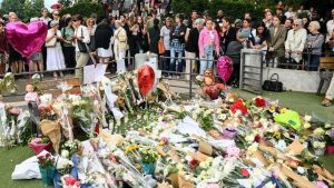 A large crowd of people gather behind a patch of grass in the Annecy park which is covered with bouquets of flowers, balloons and teddy bears.