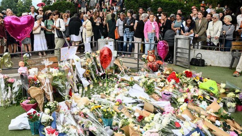 A large crowd of people gather behind a patch of grass in the Annecy park which is covered with bouquets of flowers, balloons and teddy bears.