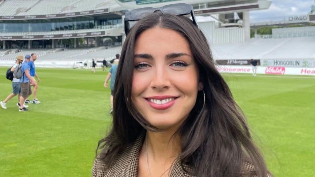 A brown hair young woman is standing on a cricket pitch. Behind her is a press stand and other seats for spectators. She is looking straight at the camera and smiling. She wears a jacket and sunglasses on her head.
