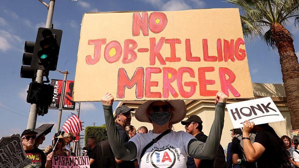 Protester holds a sign reading "No job-killing merger" in front of a crowd outside Paramount Studios in Los Angeles in September, 2026.